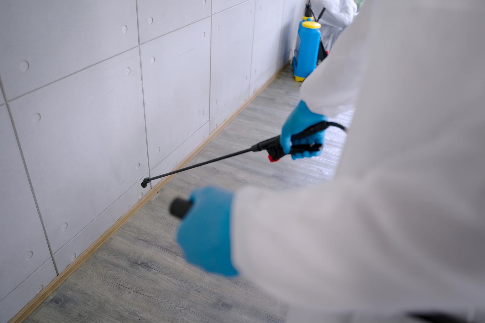 Technician in a protective suit and blue gloves spraying along the base of a tiled wall